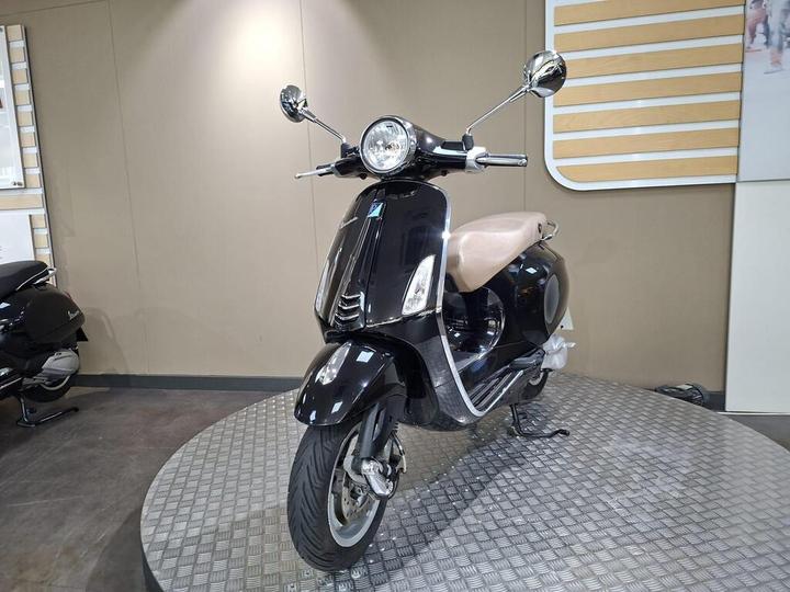 A black scooter with a beige seat is displayed on a circular platform in a showroom, featuring a chrome front with two mirrors and a round headlight. The background consists of a neutral-colored wall with wooden accents.