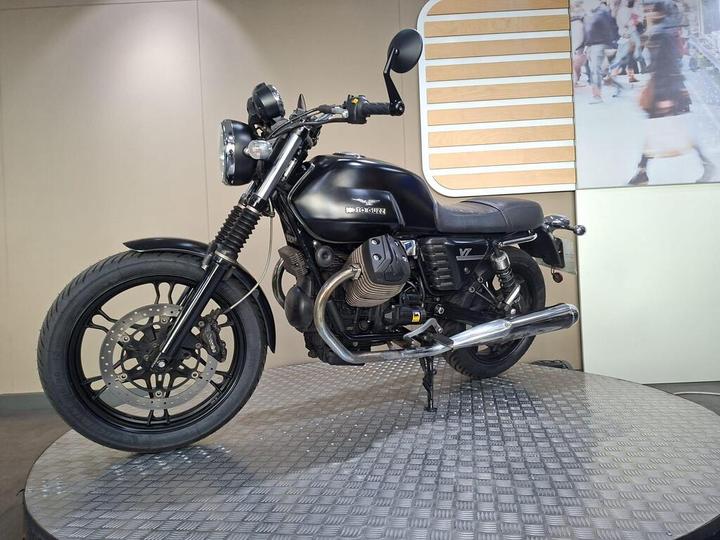 A black Moto Guzzi motorcycle is displayed on a circular platform, showcasing its sleek design and chrome details against a minimalist background.