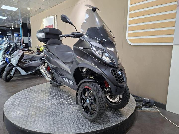 A black three-wheeled scooter displayed on a circular platform in a showroom, with a transparent windshield and sporty design, surrounded by other scooters in the background.