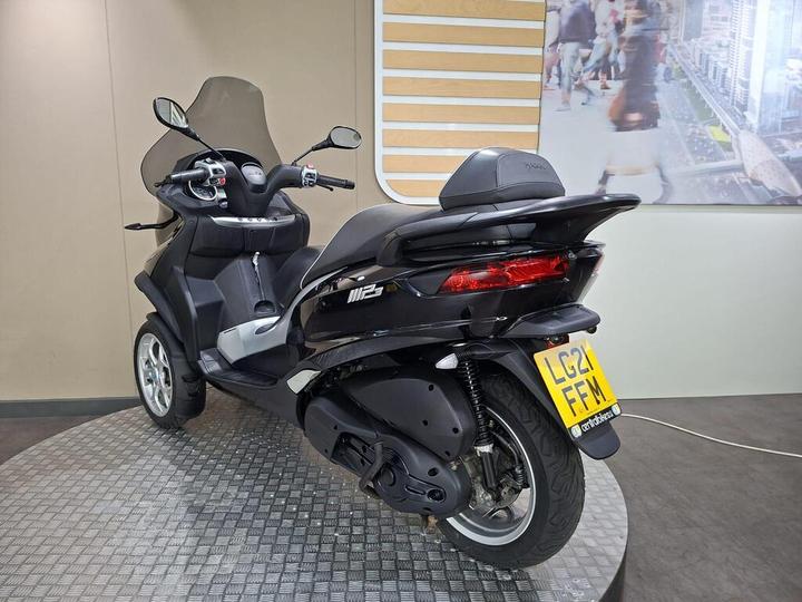 A black three-wheeled scooter with a windscreen, displayed in a modern showroom with a patterned floor and a partially visible background featuring city pedestrians and buildings. The vehicle has a UK license plate reading "LG21 FFM."