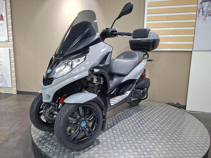 A sleek gray three-wheeled scooter with a black windshield and seat, displayed on a circular platform with a textured surface.