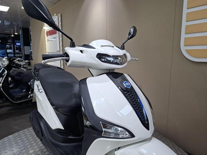 A white scooter with a sleek design, featuring a modern front display, a comfortable seat, and a mirrored handlebar, displayed in a showroom setting.