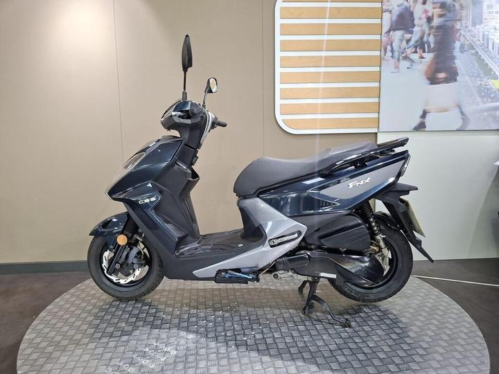 A dark blue scooter displayed on a rotating platform in an indoor setting, featuring a modern design and sleek lines. The background includes a wall with a visual display of people and cityscapes.