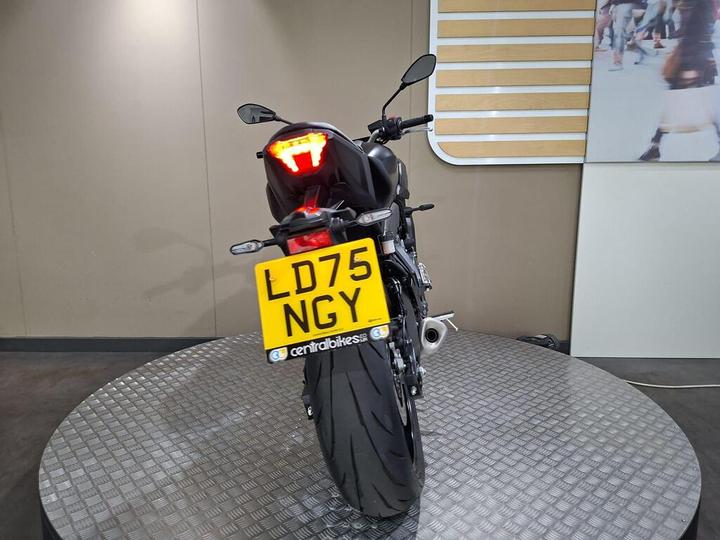 A rear view of a motorcycle displayed on a rotating platform, featuring a yellow license plate with the number "LD75 NGY." The motorcycle has a sleek design and prominent rear light. The background is a plain wall with a subtle gradient.