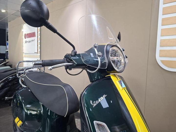 A close-up view of a dark green Vespa scooter with a yellow stripe, featuring a transparent windscreen, handlebar with mirrors, and a front headlight, displayed against a neutral background in a showroom.