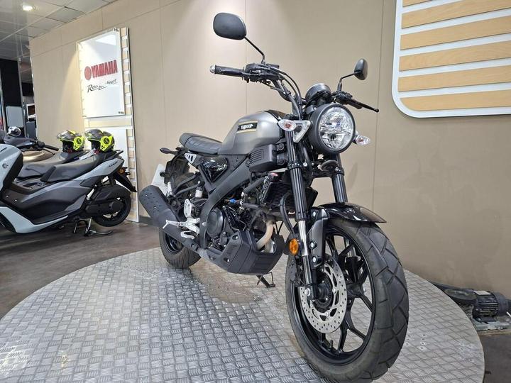 A sleek, matte black Yamaha motorcycle is prominently displayed in a showroom, with modern scooters and helmets visible in the background. The bike features a round headlight, dual disc brakes, and a distinctive design.
