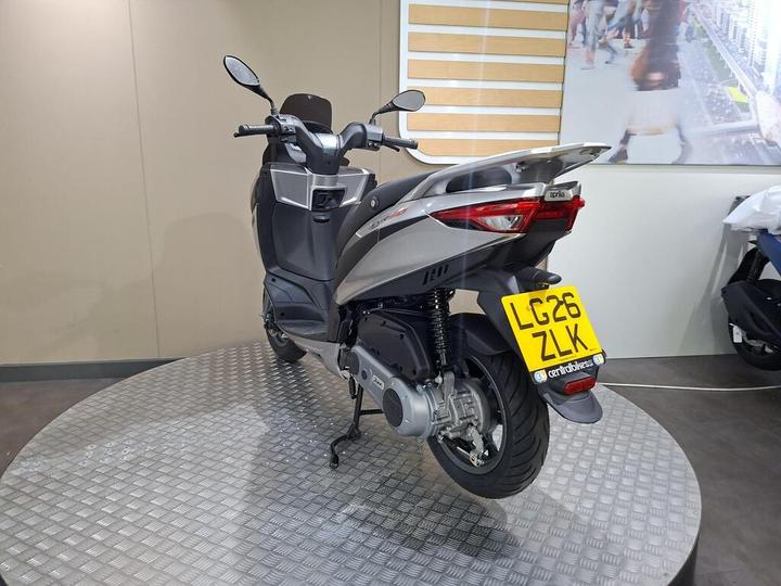 A silver scooter displayed in a showroom setting, featuring a clear view of its rear, side, and license plate. The scooter is set on a circular textured platform with a neutral background.