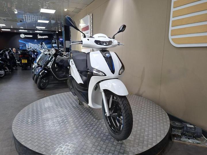 A modern white scooter displayed on a circular metal stand inside a showroom, with various other scooters visible in the background.