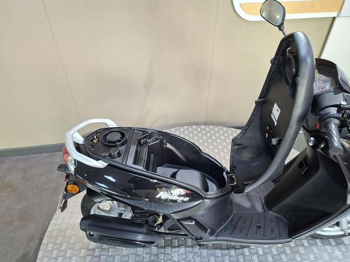Black scooter with an open seat, revealing the storage compartment and fuel tank area, set against a neutral background with a textured floor.
