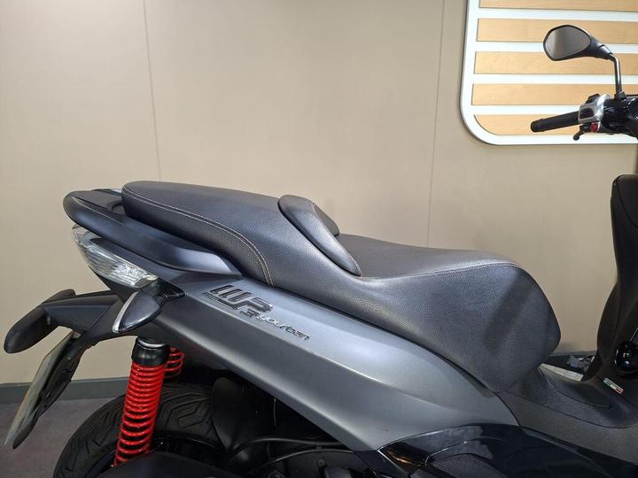 A close-up view of a motorcycle seat with a textured black cover and logo, featuring red suspension springs and part of the rear wheel visible in the frame, set against a neutral wall background.