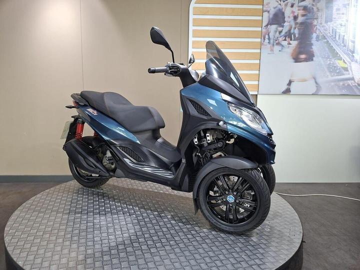 A blue three-wheeled scooter with a streamlined design is displayed on a circular platform with a textured metallic surface. The scooter features a comfortable seat, a prominent windshield, and advanced suspension visible in the rear. In the background, a blurred image of pedestrians can be seen.