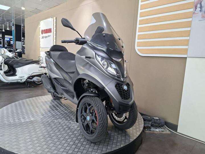 A three-wheeled scooter with a sleek gray design is displayed on a circular stand in a showroom, with a Yamaha sign visible in the background.