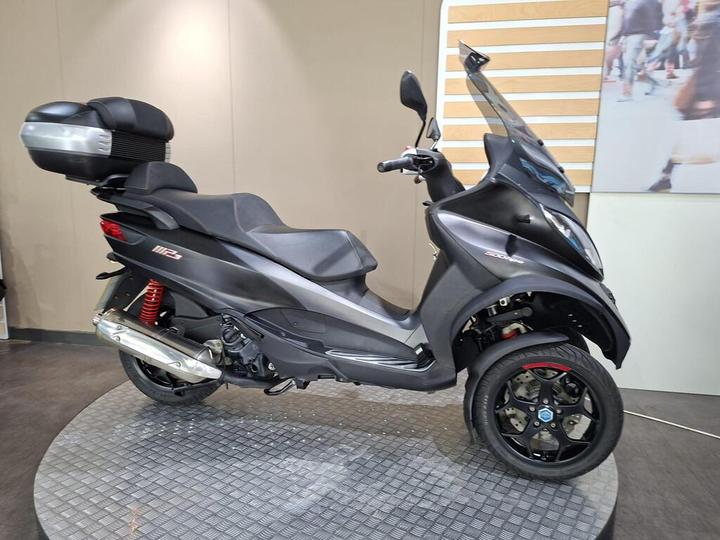 A sleek, black three-wheeled scooter displayed on a circular platform, featuring a prominent windshield, red shock absorbers, and a rear storage compartment. The backdrop includes light-colored walls and a blurred crowd scene.