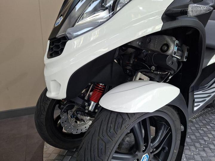 Close-up of a white scooter's front wheel and suspension, featuring a visible brake system and tire tread patterns, set against a sleek interior background.