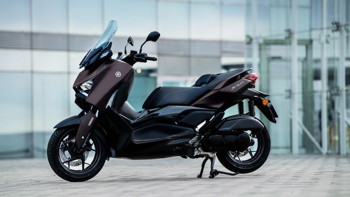 A sleek, modern Yamaha XMAX scooter in a dark finish, parked on a cobblestone surface with a glass building in the background.