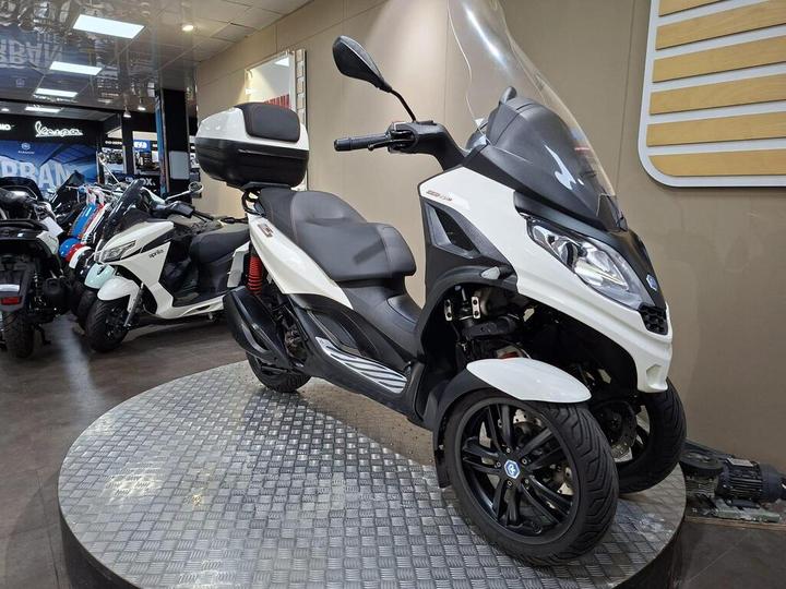 A white and black three-wheeled scooter displayed on a circular metal platform inside a motorcycle showroom, with various other scooters visible in the background.