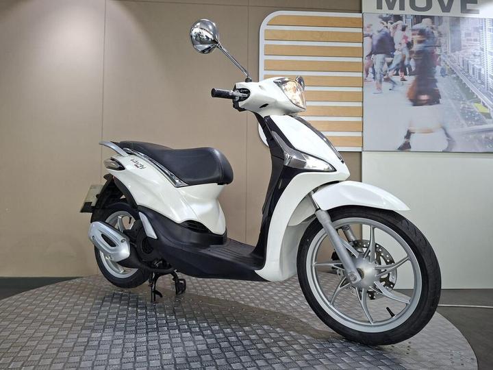 A sleek white scooter displayed on a circular metallic platform, featuring a comfortable seat and stylish design elements, with a neutral background showcasing a blurred image of pedestrians.