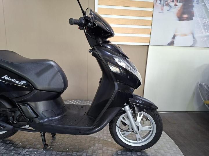 A black scooter is displayed from the side, showing its sleek design, seat, and front wheel. The background features a light-colored wall with a wooden panel and blurred figures of people.