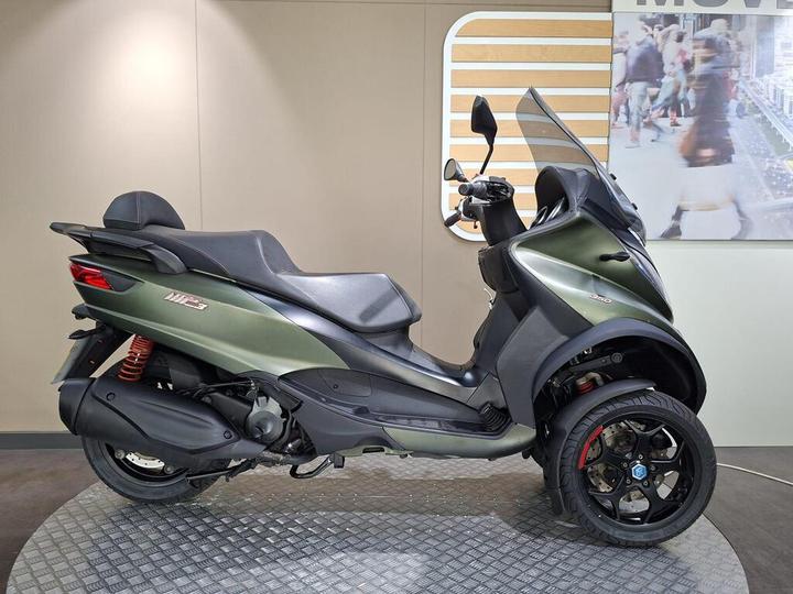 A green three-wheeled scooter displayed on a textured platform inside a showroom, featuring a streamlined design, sleek seat, and prominent rear suspension.