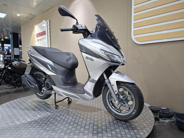 A silver Aprilia scooter is displayed on a patterned platform in a showroom, with a blurred black motorcycle visible in the background.