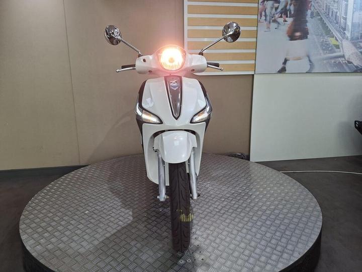 A white scooter is displayed on a circular platform, facing forward with its headlight illuminated, against a neutral background.