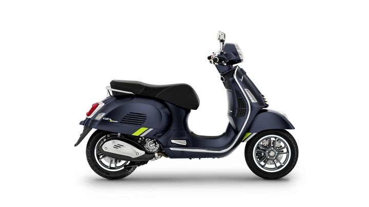 A blue Vespa scooter with green accents is displayed from the side, showcasing its sleek design, chrome wheels, and a comfortable black seat.