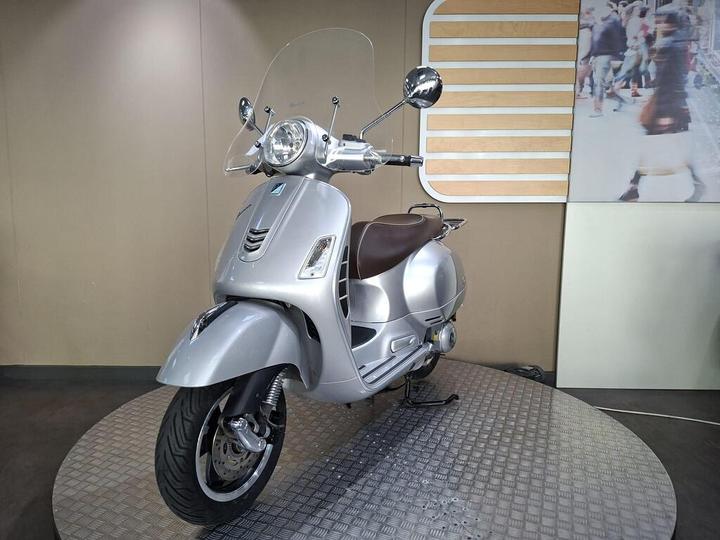A silver scooter with a retro design, featuring a clear windshield, is displayed on a circular platform. The background shows a light-colored wall with wooden panel accents and a blurred image of people walking.