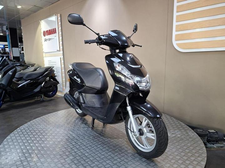 A black scooter is displayed on a circular platform inside a showroom, with other scooters visible in the background. The wall behind features a Yamaha branding sign.