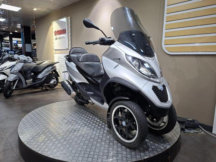 A sleek silver three-wheeled scooter displayed on a circular platform, featuring a large windshield, comfortable seating, and modern design elements, with additional scooters visible in the background.