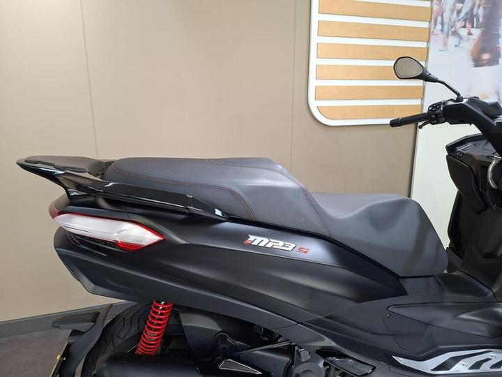 A side view of a black scooter featuring a sleek design, prominent seat, red shock absorber, and stylized rear lights, set against a neutral wall background.