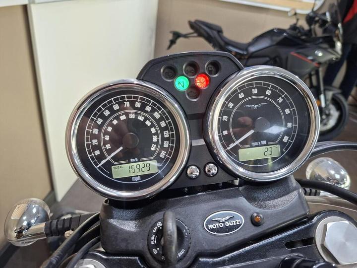 Close-up of a Moto Guzzi motorcycle dashboard displaying two analog gauges. The left gauge shows speed in km/h, with a total mileage of 15,929. The right gauge indicates RPM, with a temperature reading of 23°C. Two indicator lights are visible, one green and one red.