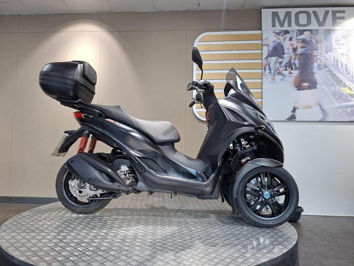 A sleek black scooter with a large rear storage compartment, displayed on a circular metal platform against a plain background, highlighting its design and features.