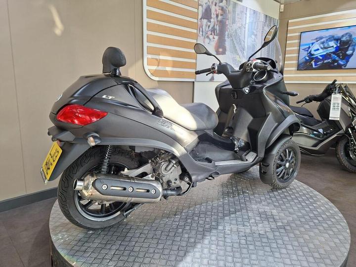 A black three-wheeled scooter displayed on a round metal platform in a showroom, featuring a modern design with a prominent rear exhaust and a license plate visible.
