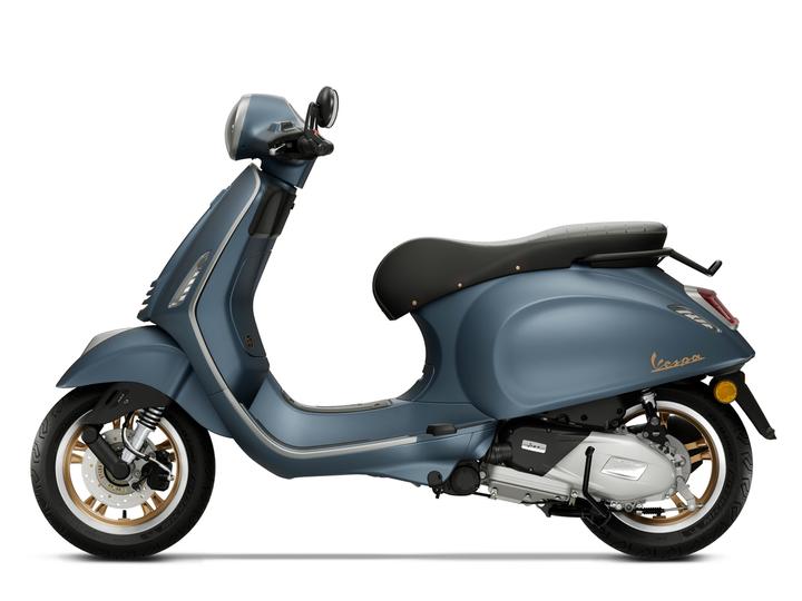 A blue Vespa scooter with a sleek design, featuring a black seat and gold accents, positioned in profile against a white background.