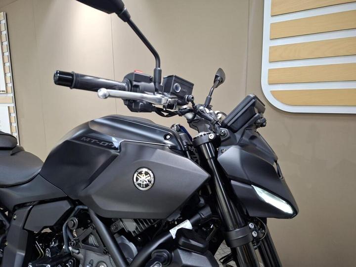 Close-up of a black Yamaha motorcycle, highlighting the handlebars, controls, and the "MT-07" model designation on the fuel tank, with a textured background.
