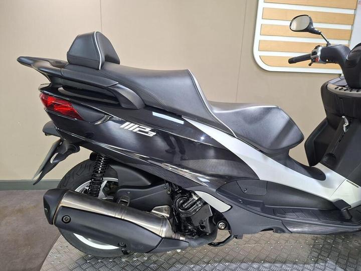 A close-up view of the rear section of a modern scooter, featuring a sleek design, black and silver color scheme, and a visible exhaust pipe. The seat is contoured, and the scooter displays the model name "P3." The background is neutral, with a simple panel design.