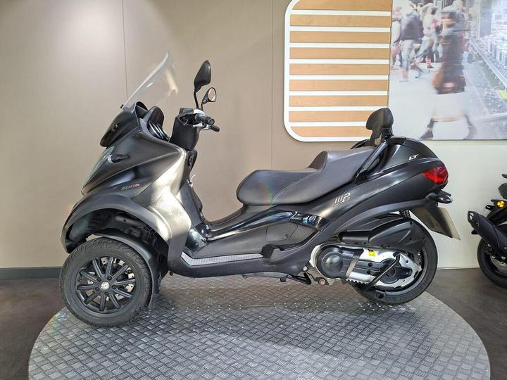 A sleek black scooter with a unique design, featuring two front wheels and a comfortable seat, displayed on a round textured platform against a neutral-colored wall.