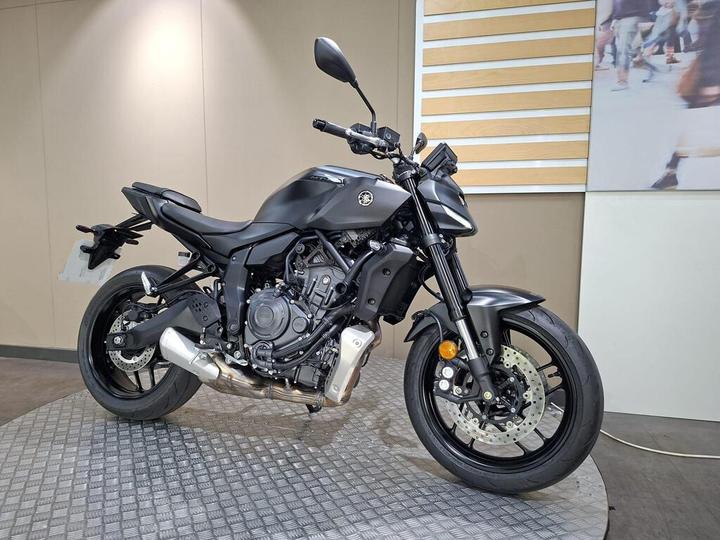 A sleek, matte black motorcycle is displayed in a showroom, showcasing its modern design and features, including a prominent engine, sporty wheels, and an upright seating position, with a neutral background highlighting its contours.