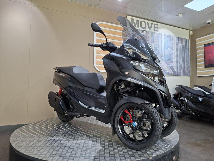 A black three-wheeled scooter with a transparent windscreen displayed on a round platform in a showroom, featuring a modern design and red accents on the wheels. The background includes a wall with a cityscape image and the word "MOVE."
