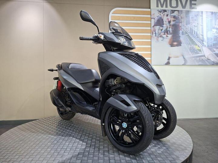 A sleek gray three-wheeled scooter displayed on a round metallic platform against a light backdrop with an image of people walking in a city.