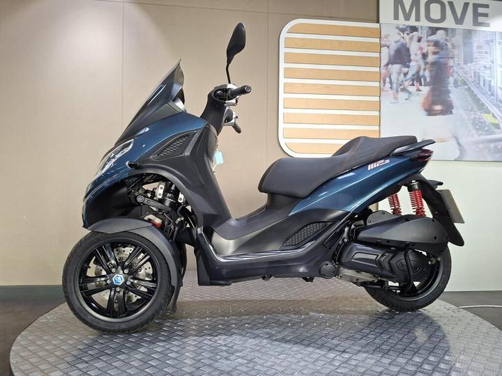 A blue three-wheeled scooter displayed on a circular platform, featuring a sleek design, front windshield, and sporty red suspension springs. The background includes a wall with a modern "MOVE" sign and a bustling street photo.
