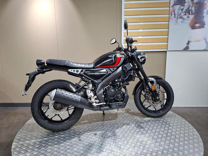 A black and red Yamaha motorcycle is displayed on a circular metal platform, set against a neutral-colored wall.