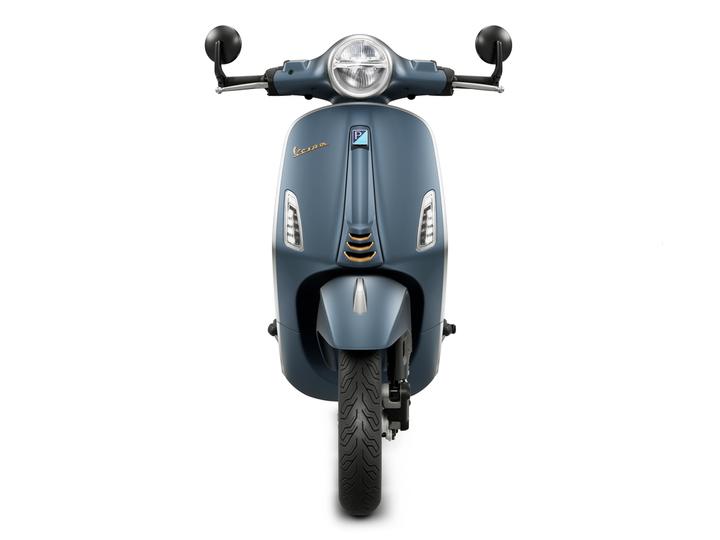 Front view of a blue Vespa scooter with a round headlight, black mirrors, and a patterned tire.