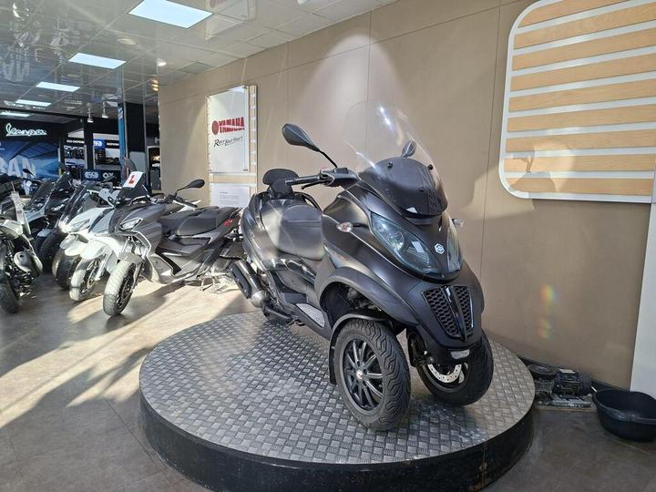 A sleek black three-wheeled scooter displayed on a circular platform in a motorcycle showroom, surrounded by other bikes and scooters. Soft lighting enhances the modern setting.