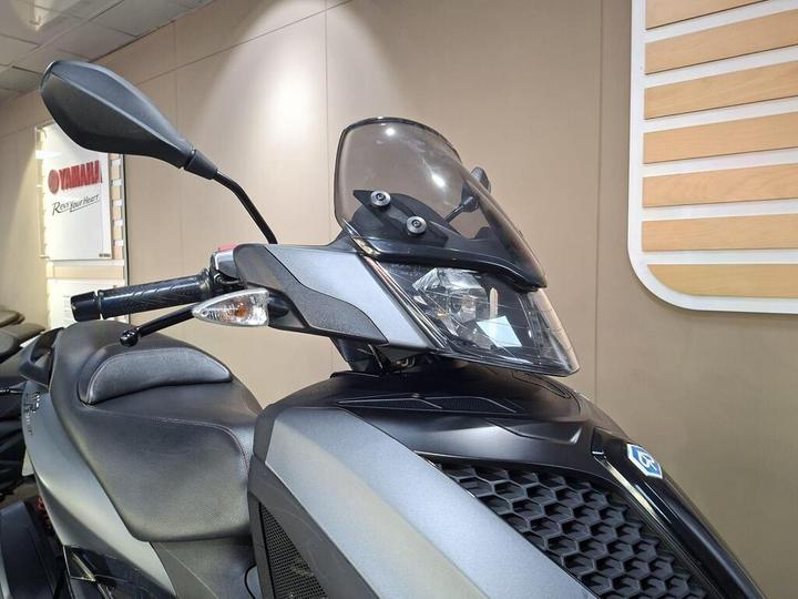 Close-up view of a gray scooter with a windshield, side mirror, and part of the seat visible, displayed in a Yamaha showroom with branding in the background.