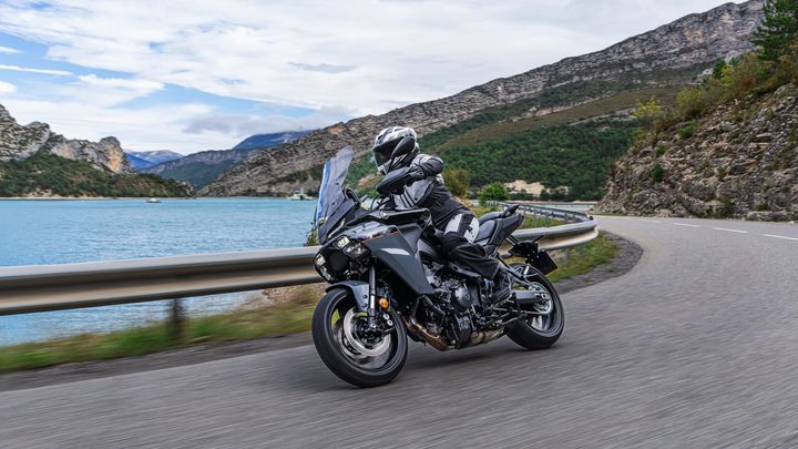 A motorcyclist in black and white gear rides a sleek touring motorcycle along a curved road beside a turquoise lake, surrounded by rocky cliffs and lush greenery under a cloudy sky.