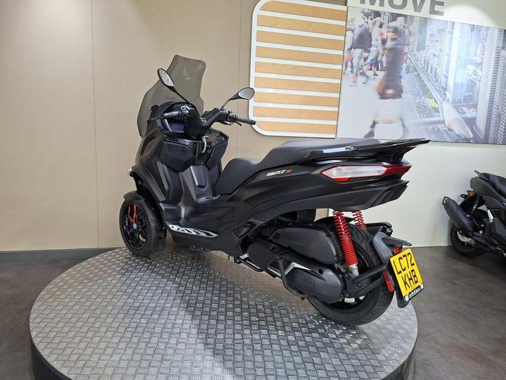 A black three-wheeled scooter is displayed on a circular metallic platform, with a clear windscreen and red shock absorbers, against a light-colored wall and a backdrop featuring a bustling city scene.