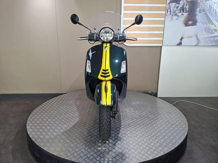 A front view of a green and yellow scooter on a circular display stand, with a transparent windscreen and chrome accents. The background features a neutral wall and a blurred image of a bustling street.