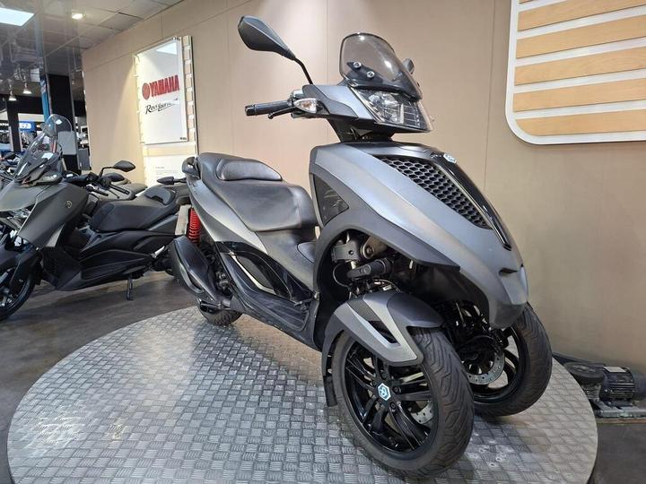 A grey three-wheeled scooter displayed on a circular platform in a Yamaha showroom, with another motorcycle partially visible in the background and signage on the wall.