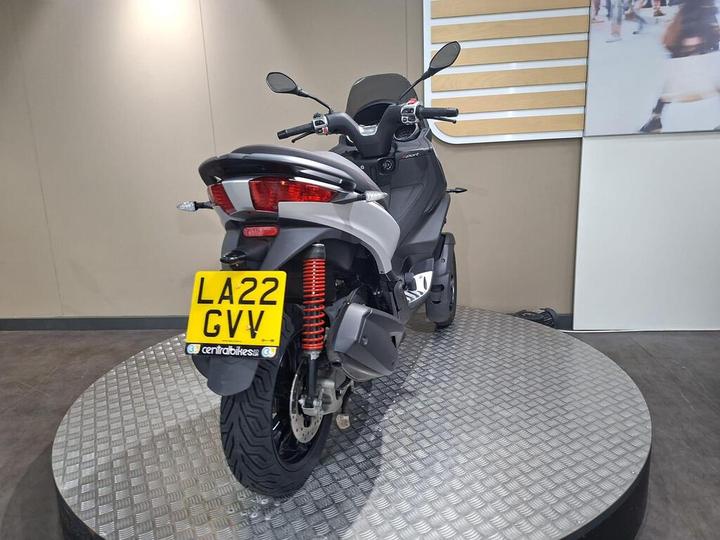 A rear view of a modern gray scooter displayed on a circular metal platform, featuring a prominent license plate reading "LA22 GV1" and showing off its sleek design and red shock absorbers.